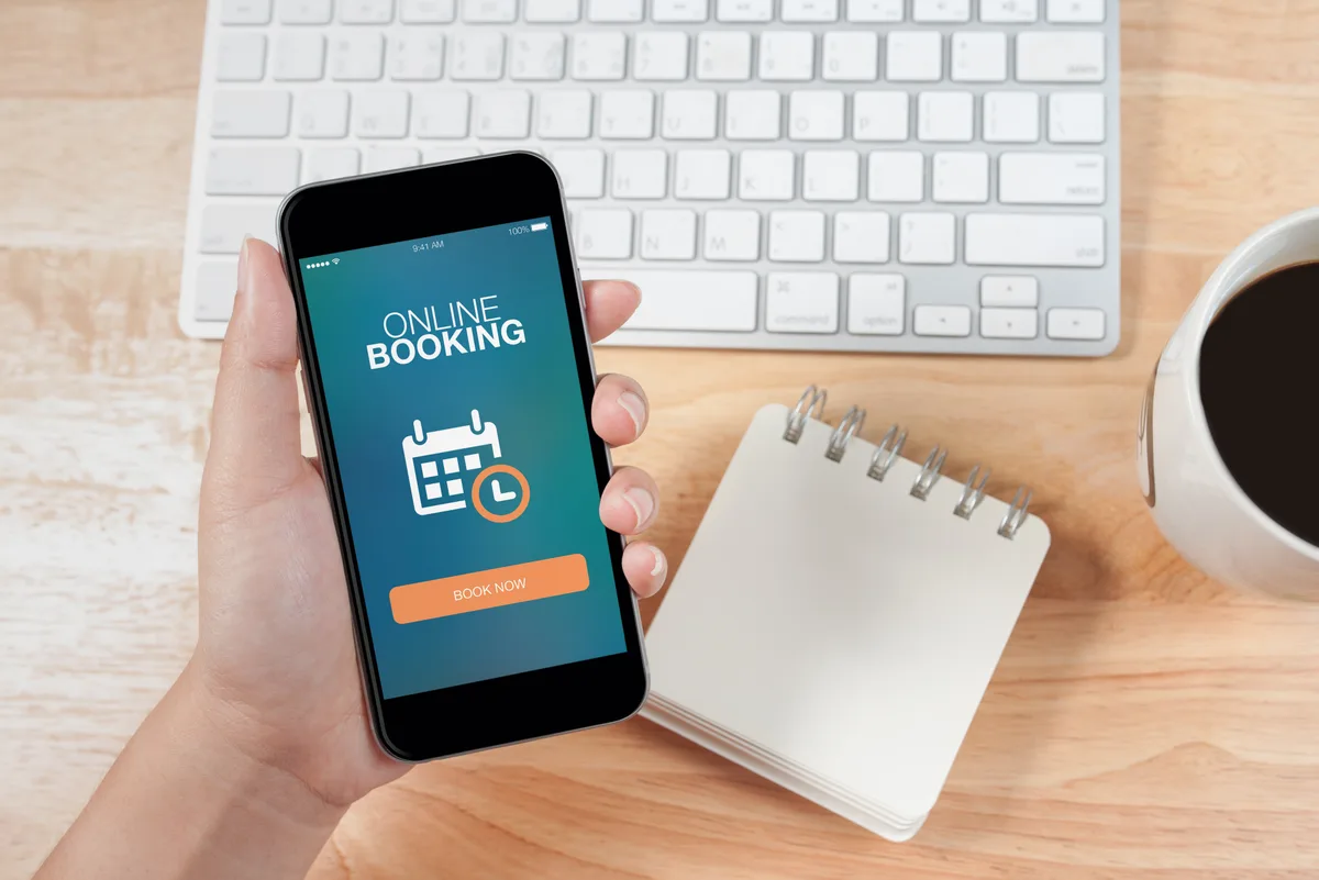 Hand holding a smartphone with an online booking app displaying a Book Now button